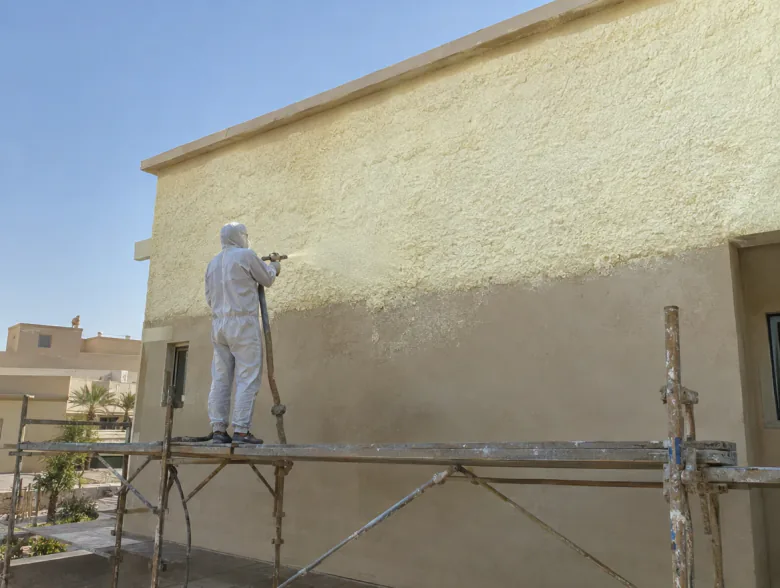 عامل يرش فوماً عازلاً على جدار فيلا خارجي نصفه مغطى